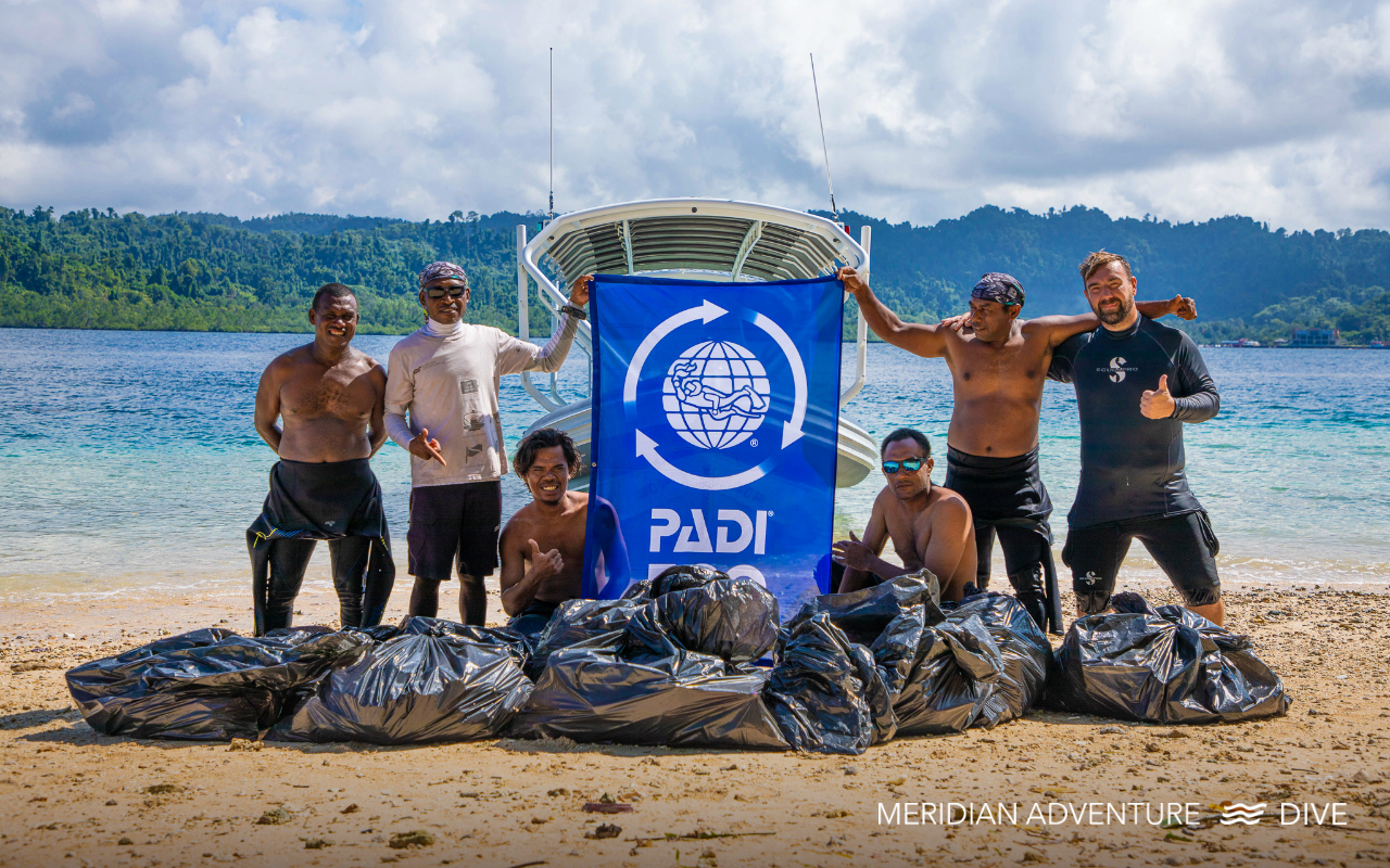 A beach cleanup done by Meridian Adventure Dive team in Raja Ampat holding a PADI AWARE banner, standing behind multiple bags of collected trash.