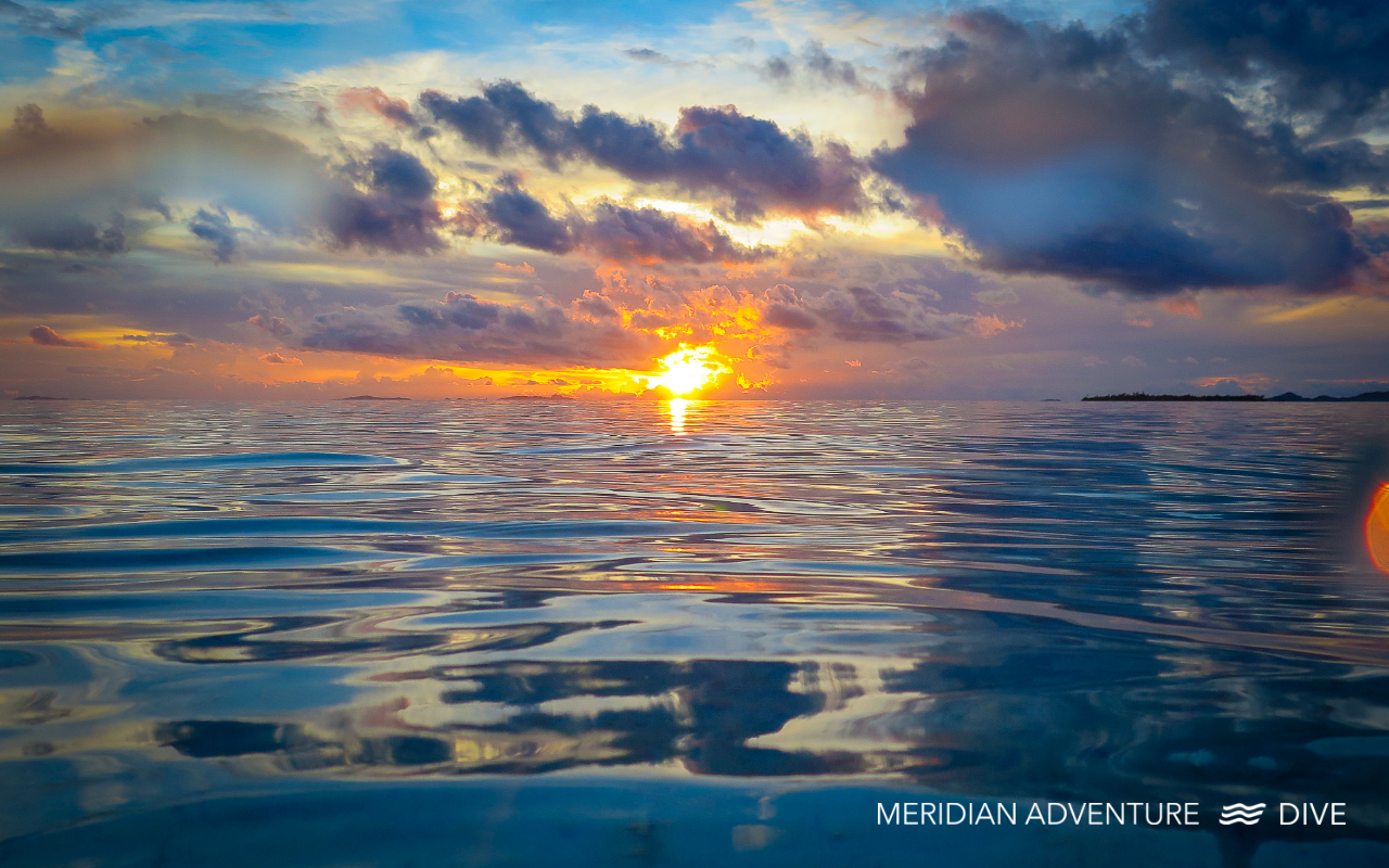 A bright sunset over calm ocean water in Raja Ampat, with orange and blue clouds reflecting on the surface.